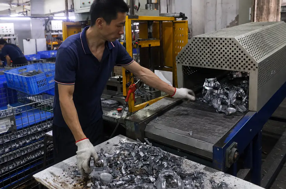 Worker handling metal components beside a conveyor and a worktable covered with parts.