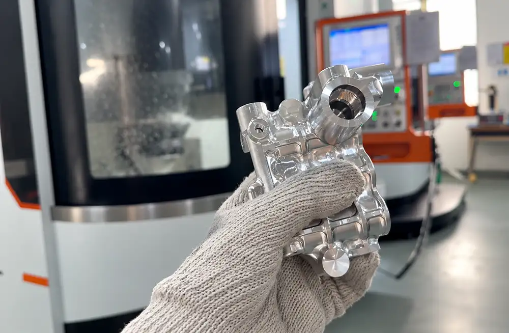 Gloved hand holding a metal component with machined pockets beside a CNC machine, illustrating CNC machining thin-walled parts.