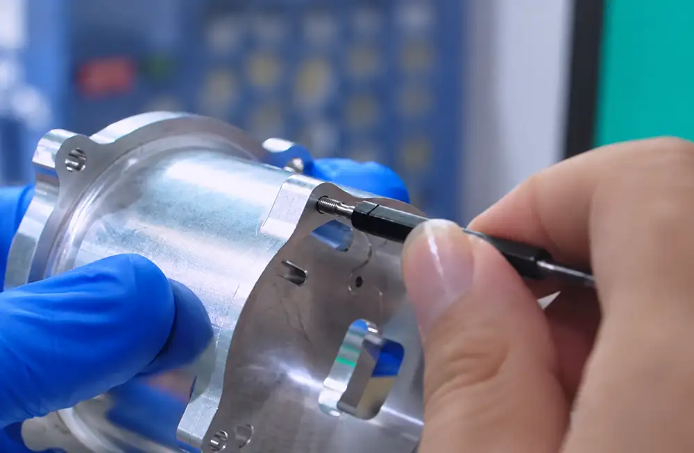 Handheld tool inserted into a threaded hole in a machined metal housing supported by a blue-gloved hand.